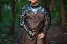 a bearded man dressed in armor stands in the woods with his hands on his hips