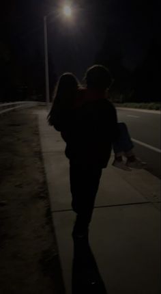 two people walking down a sidewalk at night with the street light shining on them and one person holding onto his back