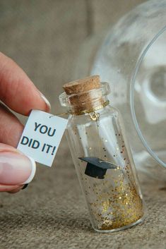 a person holding a tiny graduation cap in a glass bottle that says you did it