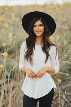 a woman standing in a field wearing a black hat and white shirt with long sleeves