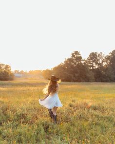 a woman in a white dress and cowboy hat walking through a field