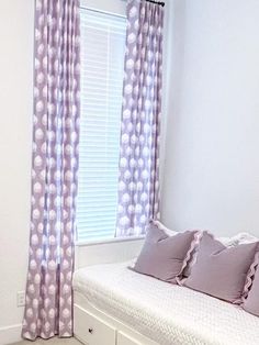 a white bed sitting next to a window with purple curtains and pillows on top of it