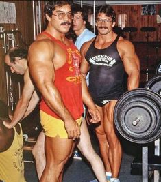 two men standing next to each other in front of a gym equipment rack with one man holding a barbell