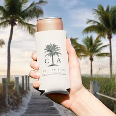 a person holding up a can cooler in front of palm trees and the ocean on a sunny day