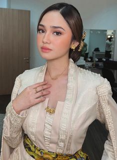 a woman in a white dress and gold jewelry posing for the camera with her hands on her chest