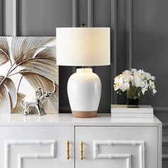 a white table lamp sitting on top of a dresser next to a vase with flowers