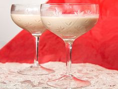 two glasses filled with liquid sitting on top of a lace covered tablecloth next to a red wall