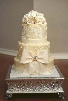 a three tiered wedding cake with white flowers and bows on the top, sitting on a silver stand