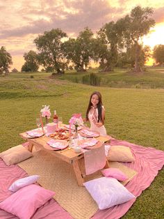 a woman sitting at a picnic table with food and drinks on it in the grass