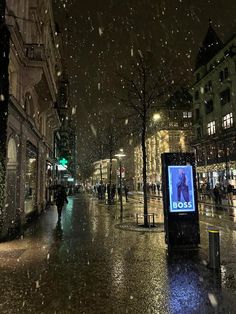 a city street at night with snow falling on the ground and people walking down the sidewalk