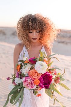 a woman with curly hair holding a bouquet of flowers in her hands on the beach