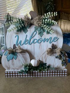 a wooden welcome sign with pumpkins and greenery on the front, sitting on a table