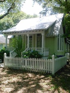 a small green house with a white picket fence and trees in the front yard on a sunny day