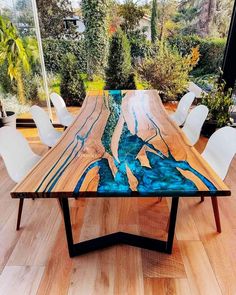 a wooden table with white chairs and blue art work on the top, in front of a large window