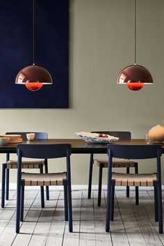 a dining room table with chairs and two pendant lights hanging over it, in front of a painting on the wall