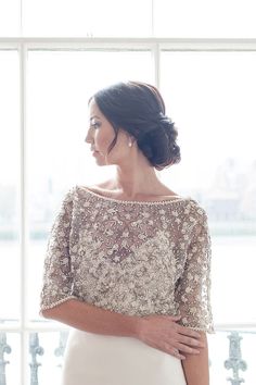 a woman standing in front of a window wearing a white dress with gold embroidery on it