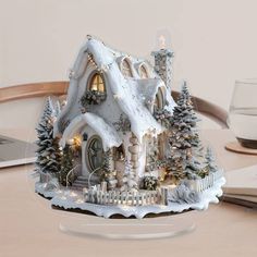 a christmas house with lights and snow on the roof is sitting on a table next to a laptop