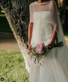 a woman in a wedding dress standing next to a tree and holding a flower bouquet