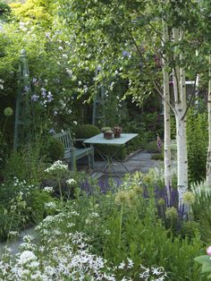 a table and chairs are in the middle of a garden with flowers, trees, and shrubs