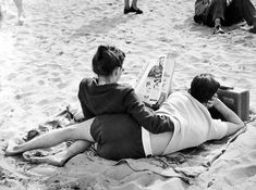 two people laying on the sand reading a book