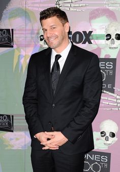 a man in a black suit and tie smiling for the camera at an event with skulls on the wall behind him