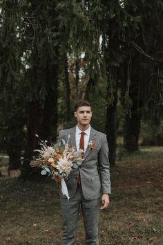 a man wearing a suit and tie holding a bouquet in front of some pine trees