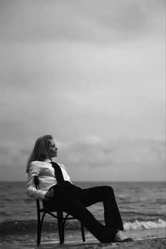 a woman sitting on top of a wooden chair next to the ocean in black and white