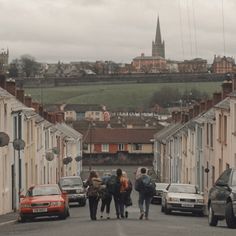 three people walking down the street in front of parked cars and buildings with a church steeple in the background