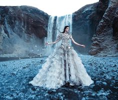 a woman standing in front of a waterfall wearing a long dress with feathers on it