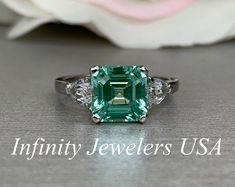 an emerald and diamond ring sitting on top of a table next to a white rose