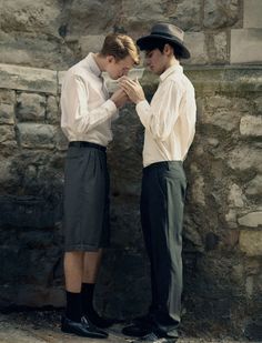 two men standing next to each other in front of a stone wall with their hands together