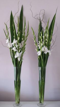 two clear vases with white flowers and greenery in them on a table against a pink wall