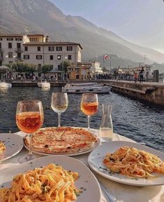 several plates of food and glasses of wine on a table near water with boats in the background