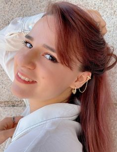 a woman with long red hair wearing a white shirt and gold hoop earrings smiling at the camera