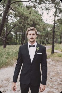 a man in a tuxedo standing on a dirt road with trees in the background