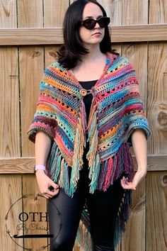 a woman standing in front of a wooden fence wearing a colorful crochet shawl