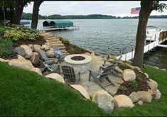 an outdoor fire pit surrounded by rocks and grass near the water with a boat dock in the background