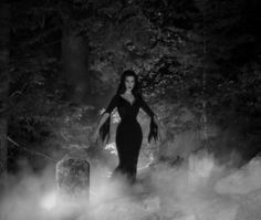 a woman in a black dress walking through the foggy woods with her hands on her hips