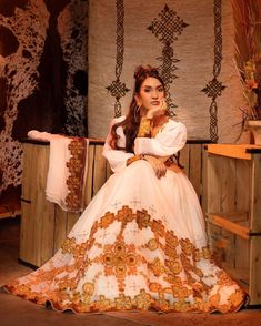 a woman sitting on top of a wooden bench wearing a white and orange dress with gold trim