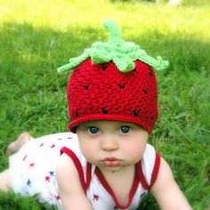 a baby wearing a crocheted strawberry hat laying in the grass with her legs crossed