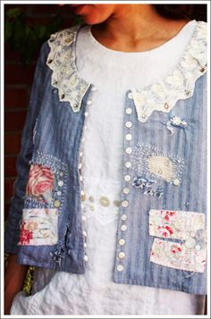 a woman wearing a blue jean jacket with patches and lace on the front, standing in front of a brick wall