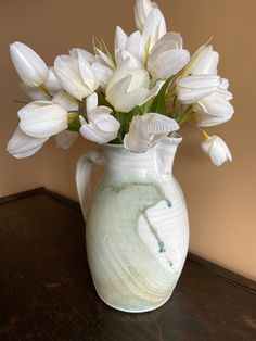 a white vase filled with lots of white flowers on top of a wooden table next to a wall