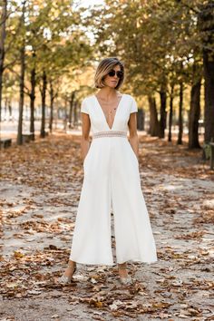 a woman in white jumpsuits and sunglasses standing on the ground with trees behind her