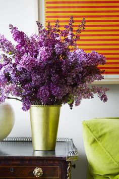 purple flowers are in a gold vase on a table next to a green chair and yellow pillow