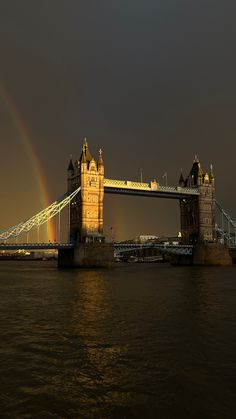 the tower bridge is lit up at night with a rainbow in the sky behind it