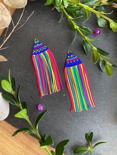 two pairs of colorful fringe earrings sitting on top of a wooden table next to green leaves