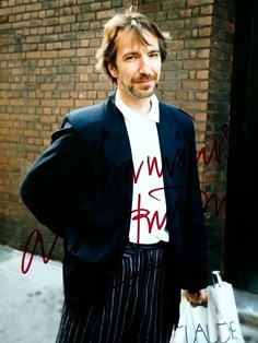 a man with long hair wearing a jacket and striped pants holding a white bag in front of a brick wall