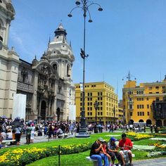 people are sitting on benches in the middle of a park with yellow buildings and green grass