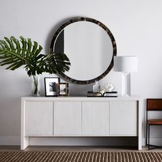 a white dresser topped with a round mirror next to a potted plant and framed pictures