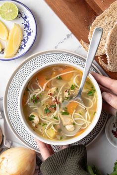 a person holding a spoon in a bowl of chicken noodle soup with lemons and parsley